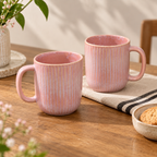 Cherry Blossom Mug Set of 2 in soft pink with textured finish with vertical fluted ridges, displayed on a wooden table with a striped cloth, a plant, and a small bowl of cookies.