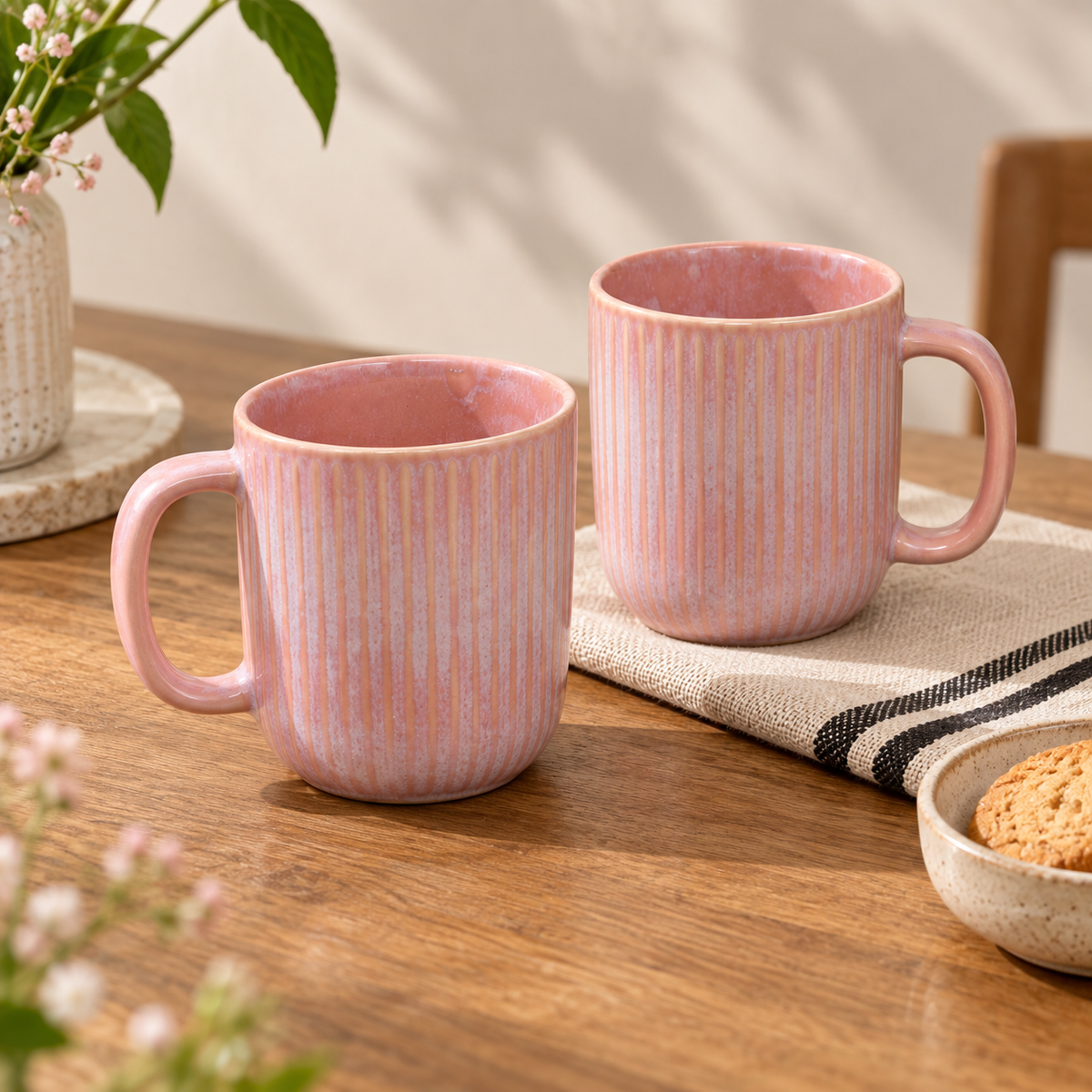 Cherry Blossom Mug Set of 2 in soft pink with textured finish with vertical fluted ridges, displayed on a wooden table with a striped cloth, a plant, and a small bowl of cookies.
