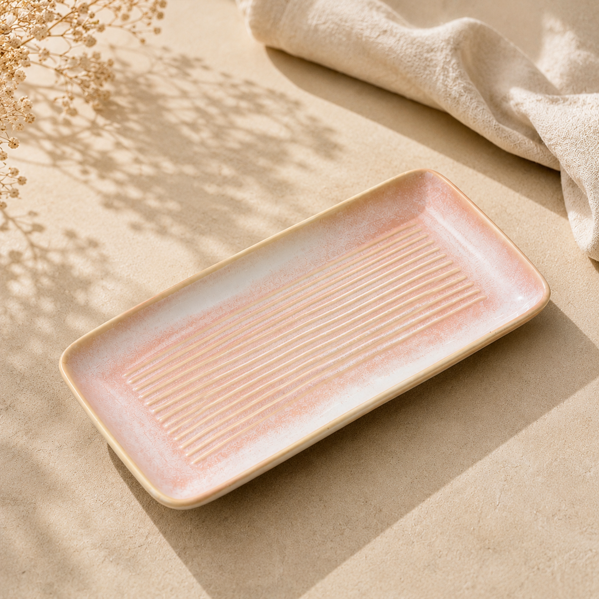 A rectangular ceramic serving tray by Ozay Living with a pale pink and beige gradient glaze, featuring a ribbed center. The tray is styled on a textured beige surface with soft shadows from dried flowers and a beige cloth in the background.