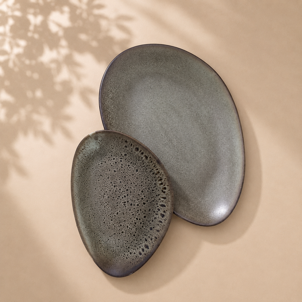 Two organic-shaped ceramic plates in a matte charcoal gray finish, one flat oval and one teardrop-shaped plate with a bubbly textured glaze, displayed on a warm beige surface with soft leaf shadows.