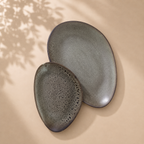 Two organic-shaped ceramic plates in a matte charcoal gray finish, one flat oval and one teardrop-shaped plate with a bubbly textured glaze, displayed on a warm beige surface with soft leaf shadows.