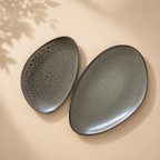 Two oval ceramic plates in a speckled dark gray glaze, one showing the textured interior and the other showing the smooth exterior, set on a warm beige surface with soft leaf shadows.