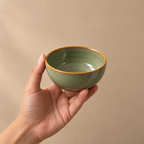 A hand holding a small ceramic bowl with a sage green glaze and a contrasting brown rim, set against a solid beige background.
