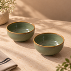 Two small glazed ceramic bowls in a muted sage green color with a rustic gold-brown rim, displayed on a beige textured tablecloth with soft natural shadows.