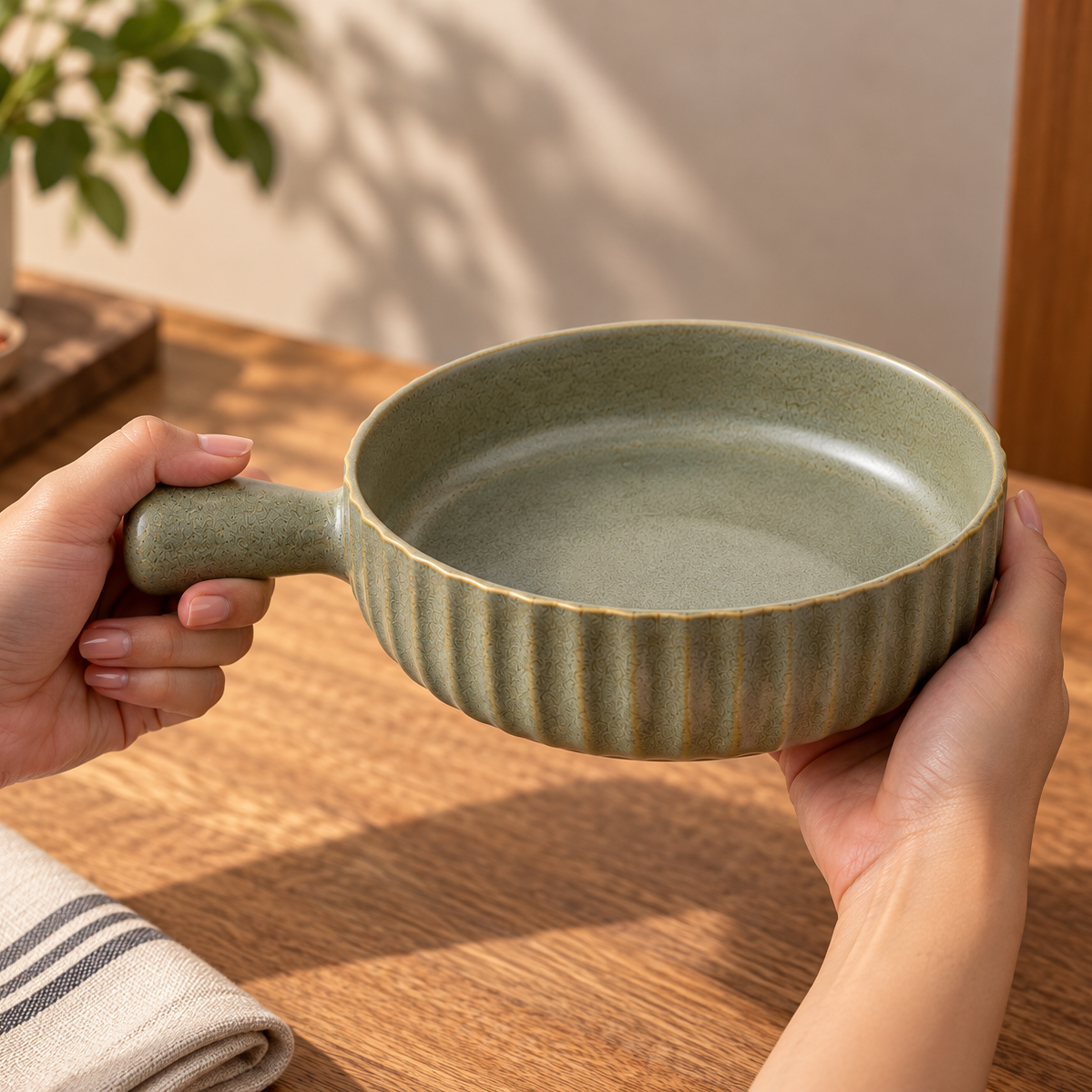 A person holds a small ceramic bowl with a fluted rim and a loop handle, shown in a warm indoor setting on a wooden table.
