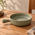 A small round ceramic pan with a handle, featuring a textured fluted exterior and a smooth matte interior, in a muted sage green color, set on a wooden table.