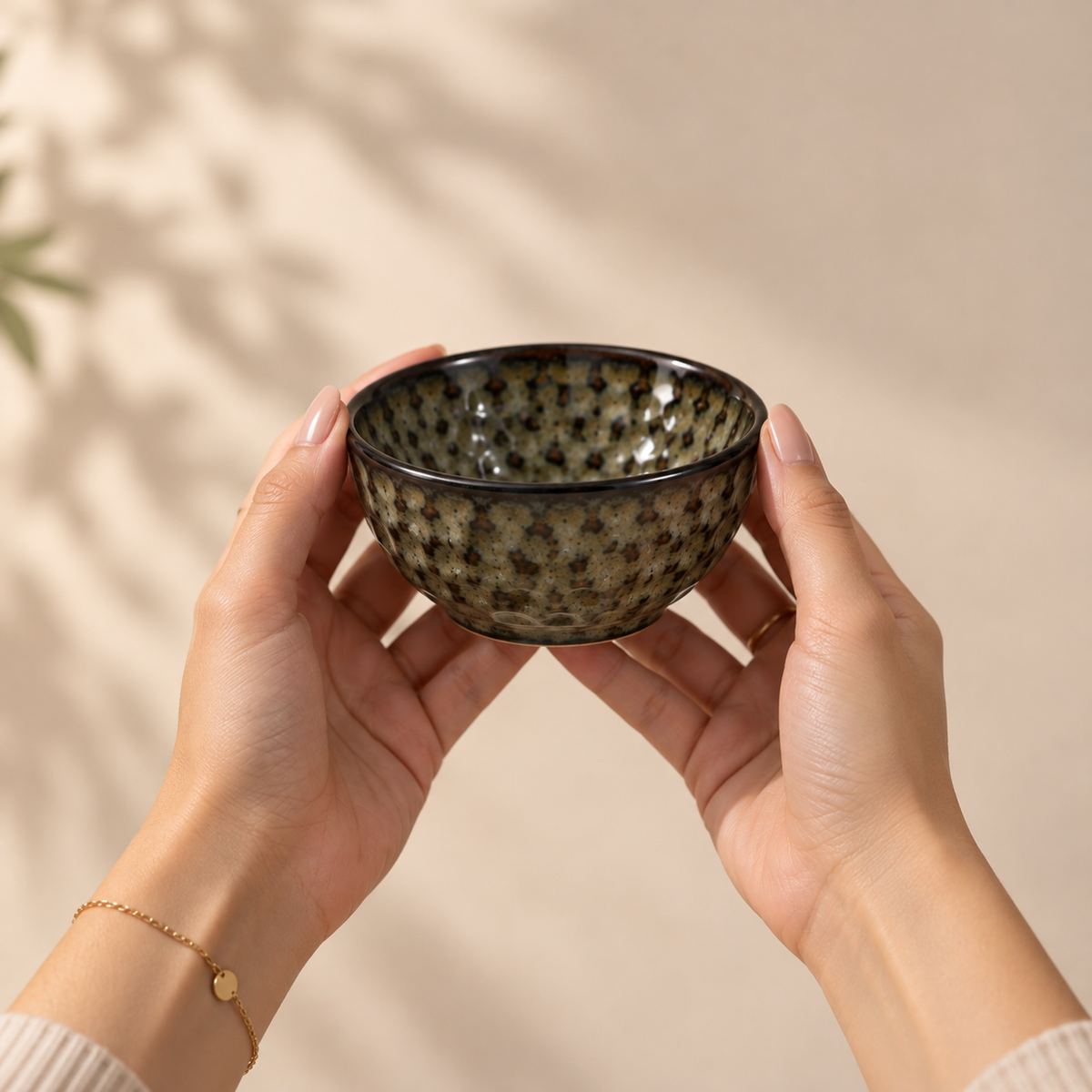 A person holds a small speckled ceramic bowl with both hands against a warm beige background with soft leaf shadows.
