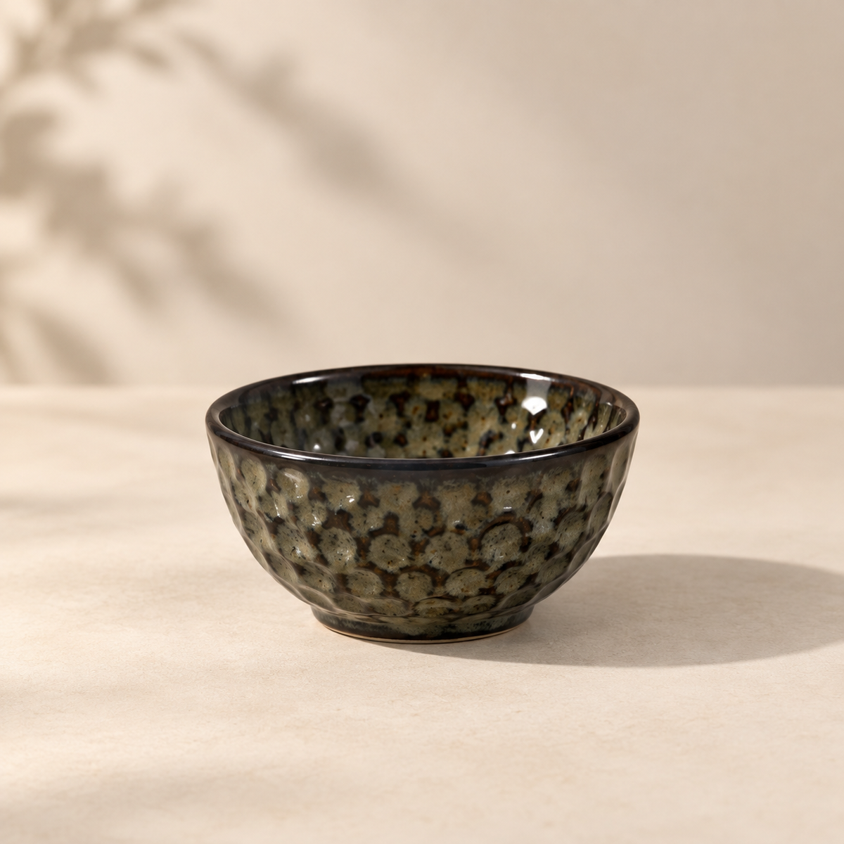 A small ceramic bowl with a textured, hammered surface and a glossy dark brown and beige speckled glaze, set on a light beige surface against a matching background with soft leaf shadows.
