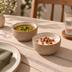 Two beige ceramic bowls on a striped table runner, one containing a yellow soup and the other a white yogurt dish with red pomegranate seeds and green herbs, in a warm, sunlit dining setting.
