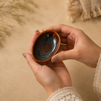 A person's hands holding a Ceramic Amber Tide Petite Server  with a glossy, mottled blue and brown glaze on a warm beige background.