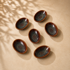 Six Ceramic Amber Tide Petite Server with a glossy dark blue-gray glaze and warm brown rims, arranged on a beige surface with soft leaf shadows.