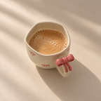 A ceramic coffee mug filled with coffee sits on a sunlit beige countertop. The mug is off-white with small red bow motifs and has a pink bow detail on the handle.