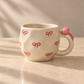 A ceramic mug with a warm cream-colored glaze, decorated with small red bow motifs and a pink bow on the handle, set on a sunlit marble countertop.