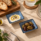 Two square ceramic appetizer dishes with a navy blue and white wave pattern, styled on a beige table. One dish contains white cheese with a drizzle of oil and herbs, and the other contains olives and dates.