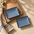 Two small square ceramic dishes with a blue and white wave pattern, styled on a wooden board and a beige cloth on a light stone surface.