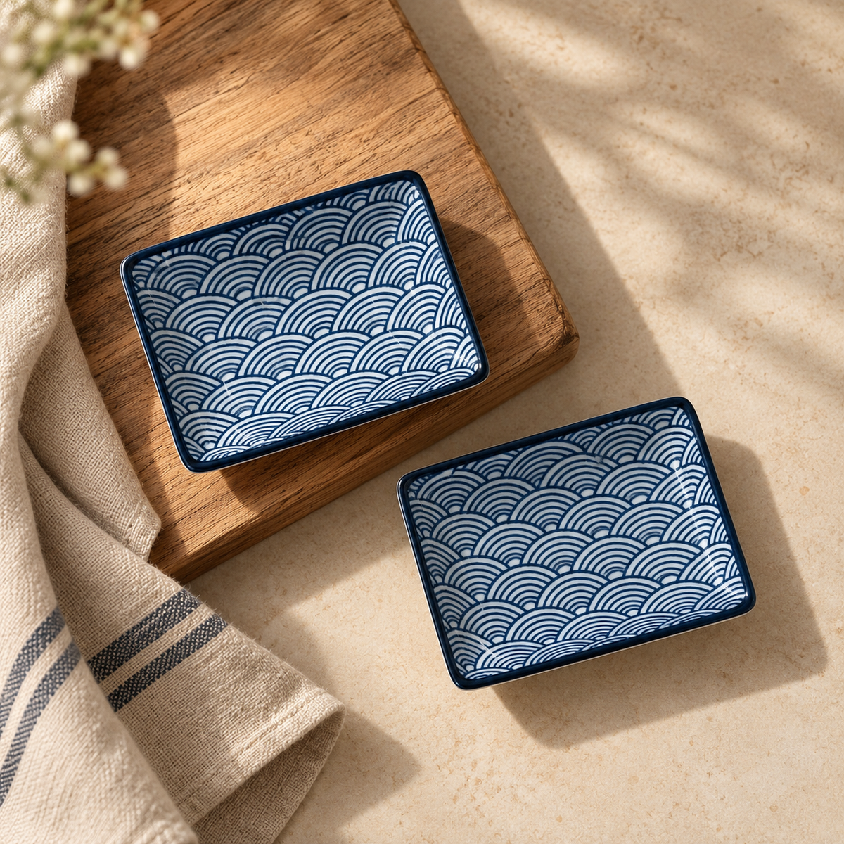 Two small square ceramic dishes with a blue and white wave pattern, styled on a wooden board and a beige cloth on a light stone surface.