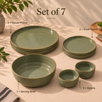 A 7-piece Grove Dinner Set dinnerware set in a muted sage green glaze with gold-toned rims, arranged on a beige tablecloth with text labels identifying each piece: 2 dinner plates, 2 small plates, 1 serving bowl, and 2 katoris. Decorative leaves and a vase appear at the edges.