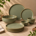 Grove Dinner Set by Ozay Living in a muted sage green glaze with thin gold trim, including plates and bowls, arranged on a beige tablecloth with soft natural shadows and decorative plants.