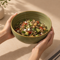 A person holds a light olive green ceramic Grove Serving Bowl filled with a colorful quinoa salad containing cucumber, tomatoes, beans, and feta cheese, set against a warm beige background.