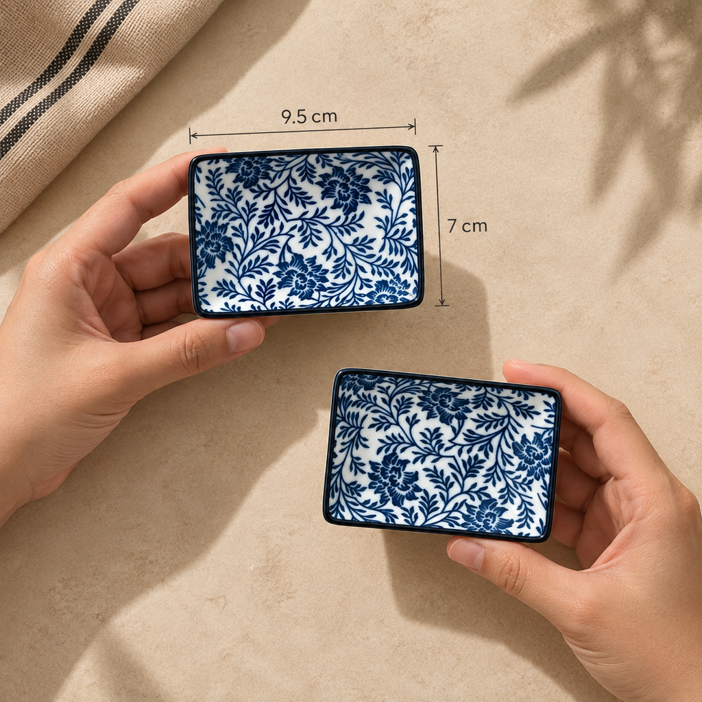 Two hands hold Indigo Bloom dip plate set of two ceramic dishes with a blue floral pattern on a white background, with dimension labels showing 9.5 cm by 7 cm, on a beige surface.