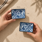 Two hands hold Indigo Bloom dip plate set of two ceramic dishes with a blue floral pattern on a white background, with dimension labels showing 9.5 cm by 7 cm, on a beige surface.