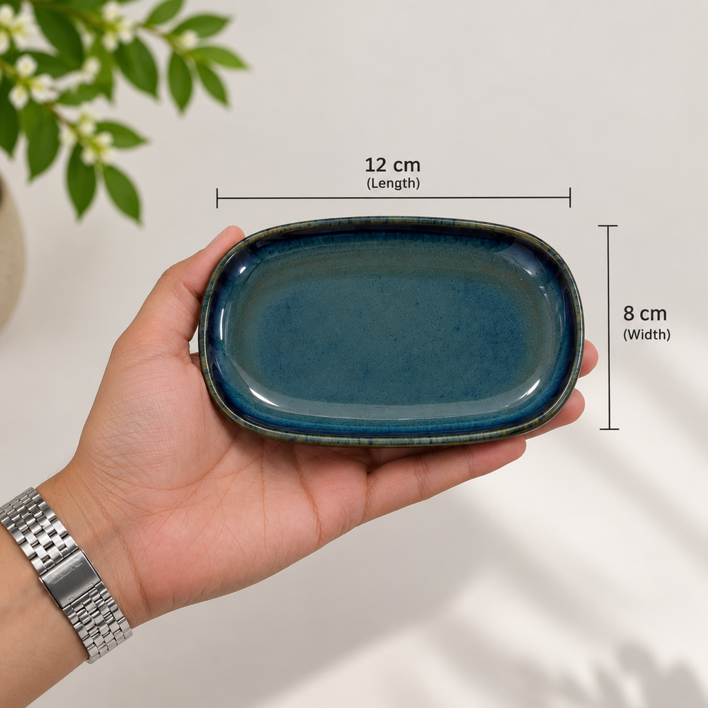 A hand holds Mira ceramic petite server with blue glaze, overlaid with dimension lines showing 12 cm length and 8 cm width, against a light background with a plant leaf in the corner.