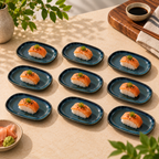 Nine Mira ceramic petite servers sushi plates arranged on a beige tabletop, each holding a piece of salmon nigiri with green garnish. A small bowl of wasabi, chopsticks, and a vase with greenery are visible around the plates in a styled dining setting.