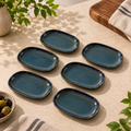 Mira ceramic petite servers in a dark teal-blue glaze with a rustic brown rim, arranged on a beige stone surface with decorative plants, a wooden board, and a striped cloth.