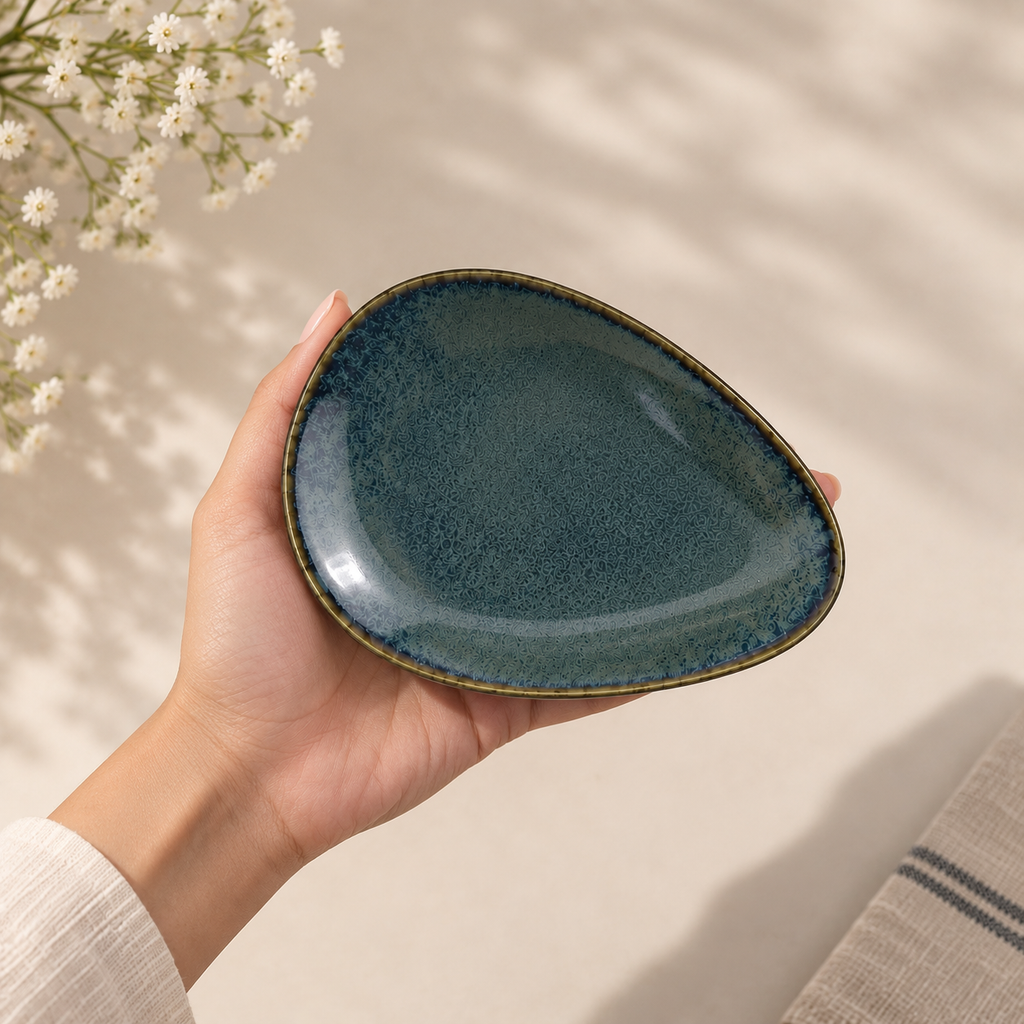A hand holding Onda Petite Ceramic Server Plate  with a speckled dark teal-blue glaze and a thin gold-toned rim, set against a softly lit neutral background with delicate white flowers in the corner.