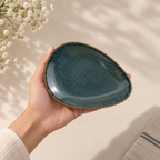 A hand holding Onda Petite Ceramic Server Plate  with a speckled dark teal-blue glaze and a thin gold-toned rim, set against a softly lit neutral background with delicate white flowers in the corner.