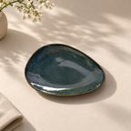 Onda Petite Ceramic Server Plate with a glossy dark teal-blue glaze and a thin gold-toned rim, styled on a warm beige surface with soft leaf shadows and small white flowers in the corner.