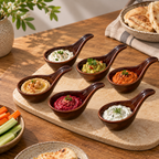 OZAY Living Scoop Dip Cups  arranged on a light stone platter, each filled with different dip or spread: hummus, guacamole, orange salsa, red baba ganoush, tzorax, and white yogurt. The platter sits on a wooden table with bread, carrot sticks, and a plant in the background.