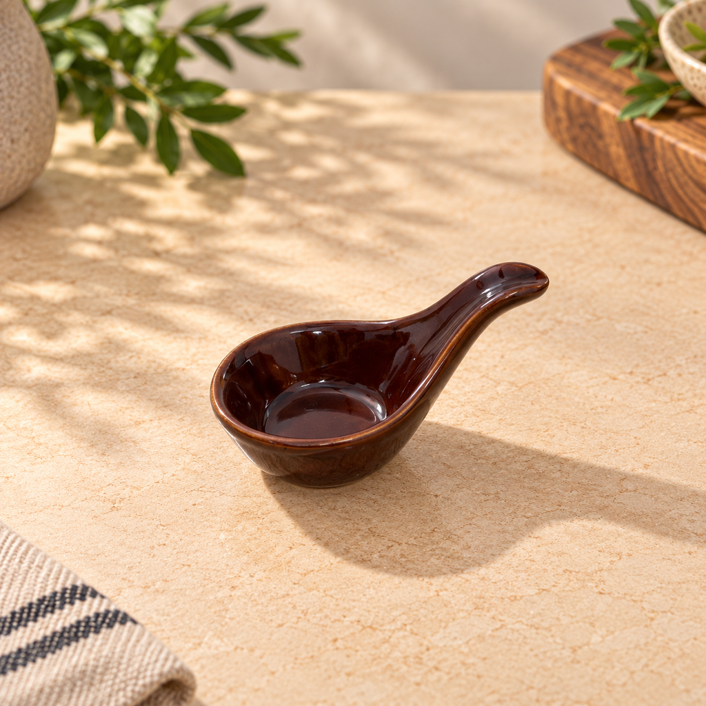A small glossy dark brown ceramic oil drizzle spoon with a handle, styled on a beige textured tabletop with soft leaf shadows.
