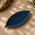 A leaf-shaped ceramic serving dish with a textured ribbed pattern, shown in a deep blue-green glaze on a beige surface with soft dappled shadows.