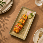 A rectangular ceramic plate with a warm beige-green glaze, styled on a wooden dining table. The plate holds grilled paneer tikka skewers with green peppers, red onions, a lime wedge, and cilantro, alongside sliced red onion.