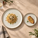 Siena ceramic plates with beige rims are styled on a warm beige stone surface. The larger round plate holds spaghetti with herbs and cheese, and the smaller oval plate holds a slice of garlic bread.