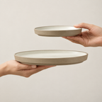 Two hands holding Siena ceramic plates  against a plain light beige background. The lower plate is larger and held horizontally by a left hand, while a smaller matching plate is held vertically by a right hand above it.