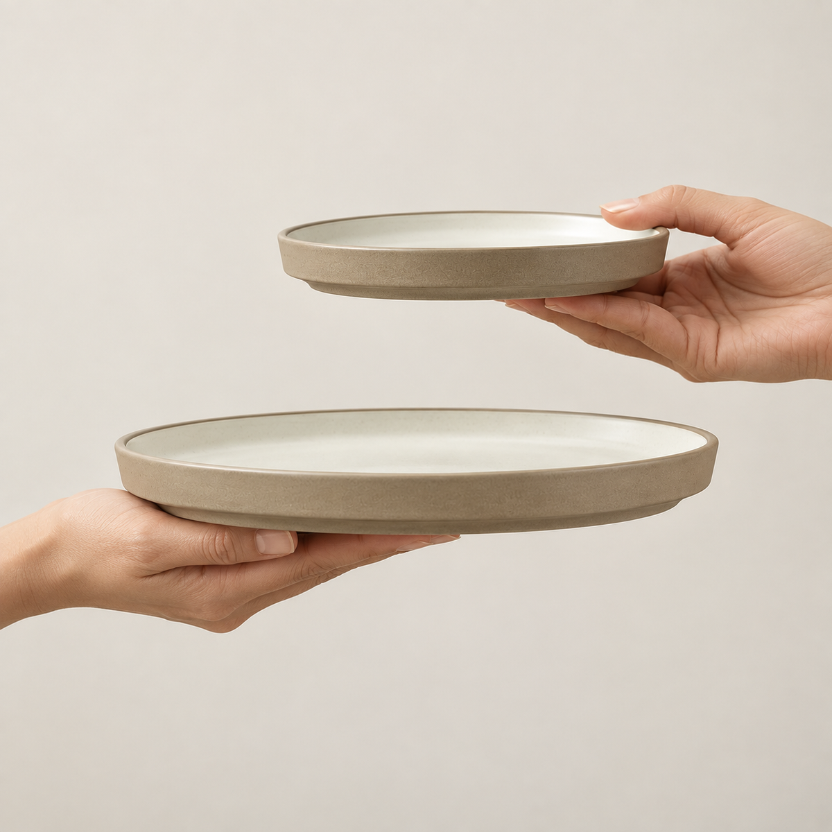 Two hands holding Siena ceramic plates  against a plain light beige background. The lower plate is larger and held horizontally by a left hand, while a smaller matching plate is held vertically by a right hand above it.