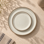 Siena ceramic plates, one larger and one smaller, stacked flat-lay style on a textured beige stone surface with soft leaf shadows, white flowers, and a striped napkin.